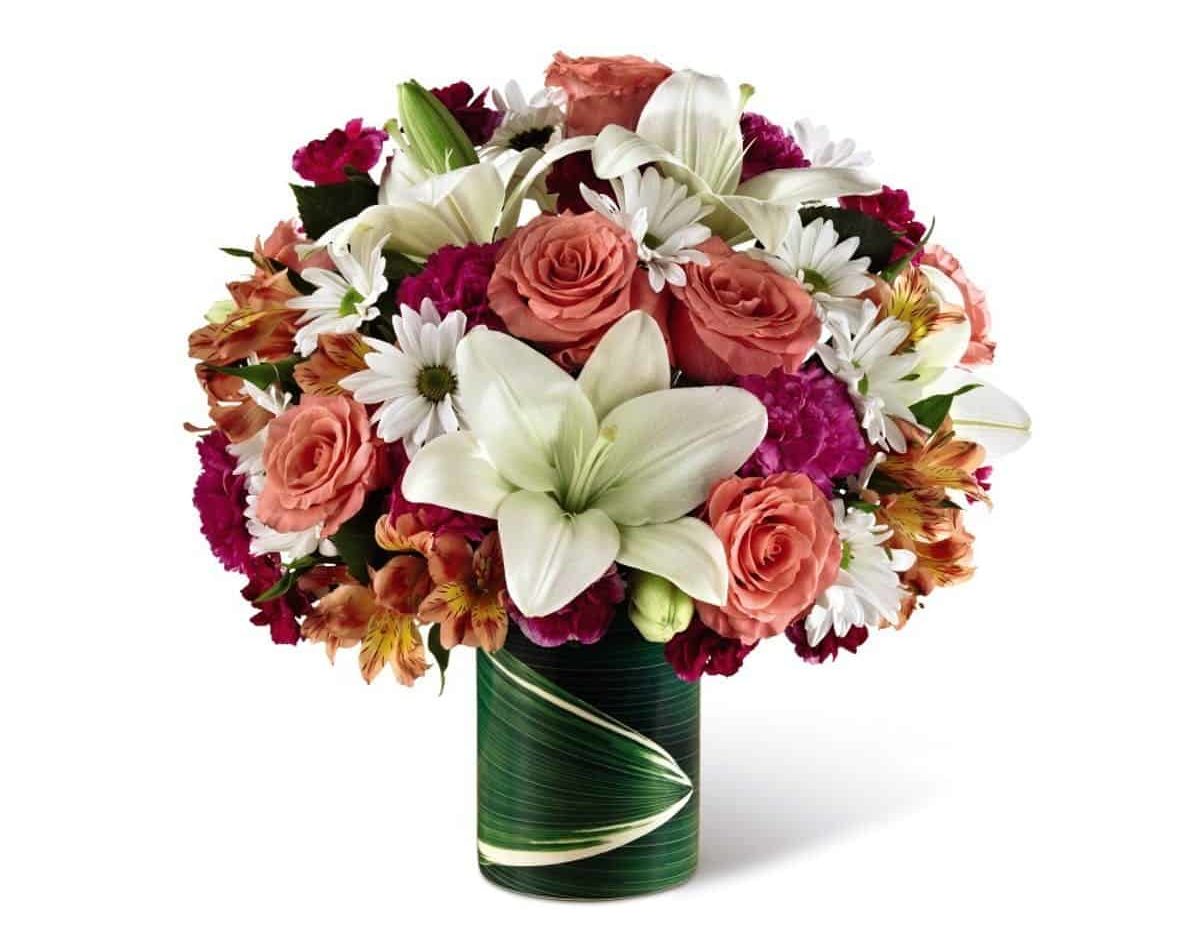 Lavender carnations, white Asiatic lilies and daisy poms, orange Peruvian lilies, coral roses (in Deluxe, Premium and Exquisite versions) and more are arranged in a glass vase.